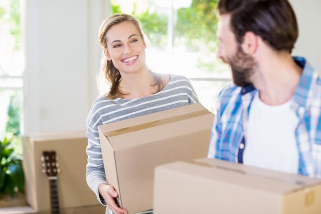 This image shows a happy young couple holding cardboard boxes while moving into their new home. The bright and airy room, along with their smiles, suggests excitement and positivity. Ideal for use in real estate, moving services, home improvement, and lifestyle blogs or advertisements.