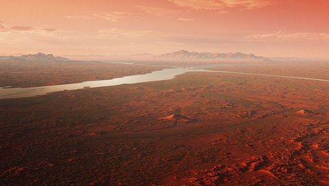 Rust-red desert river at sunset, meandering waterway carving badlands and distant peaks