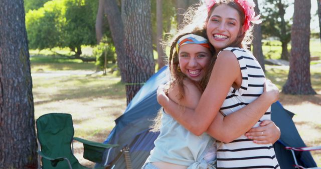 This image depicts two happy young friends hugging at a campsite with tents and trees in the background. Ideal for depicting friendship, outdoor activities, camping trips, summer adventures, and joyful moments. Perfect for use in travel promotions, camping gear advertisements, lifestyle blogs, social media posts about outdoor activities, and health and wellness websites.
