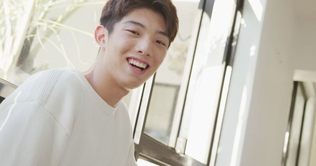 Young man smiling brightly in a well-lit room with natural sunlight. Perfect for marketing materials promoting positivity, youth lifestyles, and casual indoor settings, as well as advertisements for clothing or lifestyle brands.