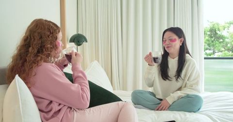 Two women relaxing on bed with pink eye patches holding glass mugs cozy self-care night