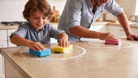 Smiling father and son cleaning kitchen countertop with colorful sponges and soap suds