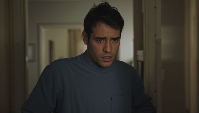 Worried man standing in dim hallway wearing blue high-neck shirt, conveying tension