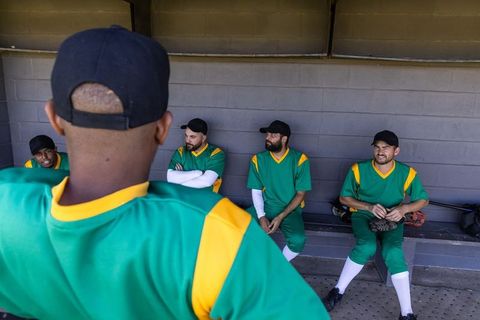 Diverse baseball teammates sitting in dugout wearing green uniforms holding gloves