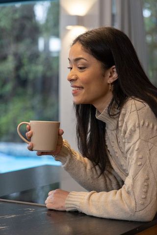 Woman holding ceramic mug by window wearing cozy cream sweater enjoying serene morning