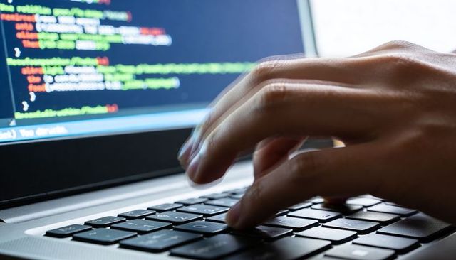 Close-up right hand typing on laptop keyboard with colorful code on screen