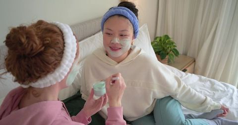 Friends applying green facial mask while relaxing on bed wearing cozy loungewear together