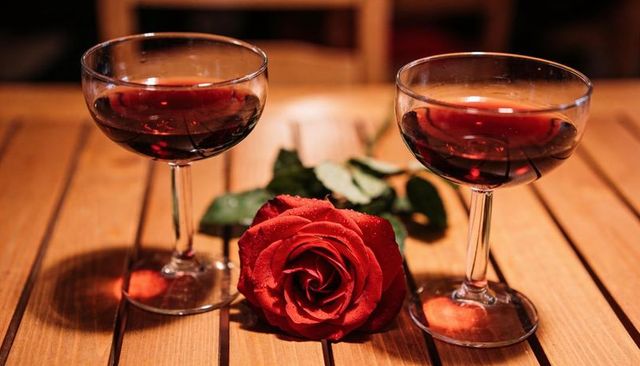 Resting red rose draped between two coupe glasses holding red wine on warm wooden table