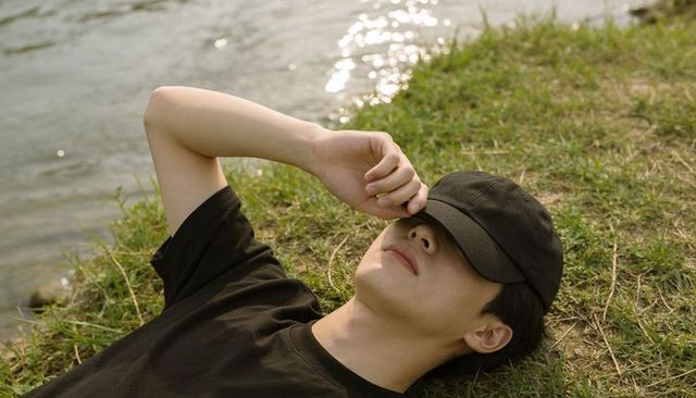 Young man reclining on grassy riverbank shielding eyes with cap, sunlit serene moment