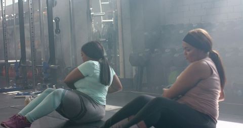 Diverse women doing seated Russian twists on mats in gym with dumbbell rack and sunlight
