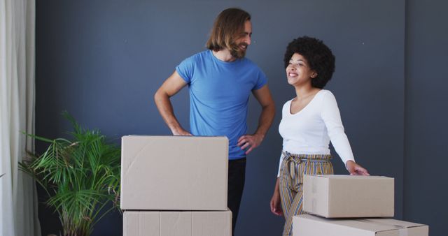 This image depicts a cheerful biracial couple standing amid stacks of cardboard boxes in their new apartment, symbolizing a fresh start. Both appear joyous and upbeat, likely on the day they moved into their new home. Their stance and the scattered boxes convey an exhilarating start to a new chapter. This photo is ideal for use in articles or advertisements about moving services, real estate promotions, new homeowners, and content focused on living spaces or milestones in life.