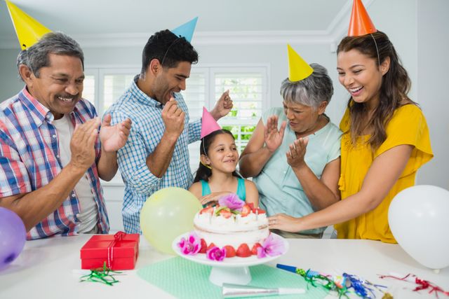 Multigenerational family celebrating a birthday at home, featuring grandparents, parents, and a child. They are wearing party hats, clapping, and smiling around a decorated cake with balloons and presents. Ideal for use in advertisements, family-oriented content, and celebration-themed promotions.