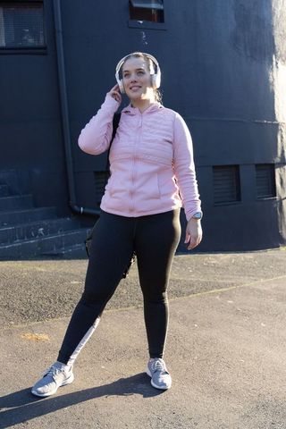 Woman listening to music on wireless headphones during urban walk wearing pink jacket