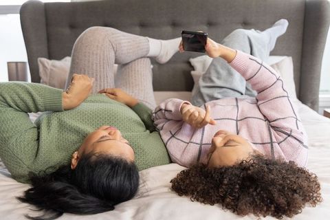 Diverse women lounging on bed in knit sweaters sharing smartphone and relaxing