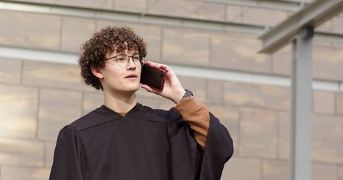 Young graduate calling on smartphone in academic gown with round glasses and curly hair