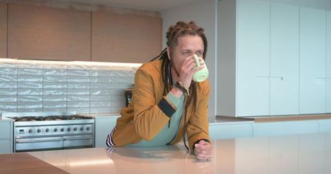 African american man sipping coffee at modern kitchen island wearing mustard jacket