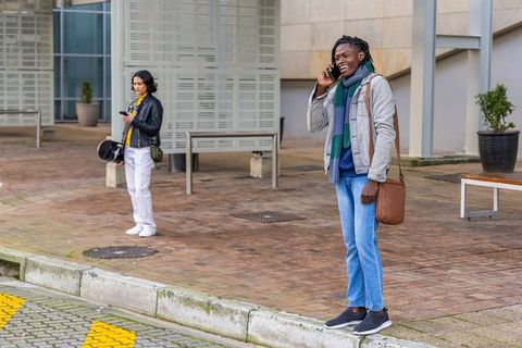 Smiling commuter talking on phone on curb with helmet-carrying pedestrian waiting nearby
