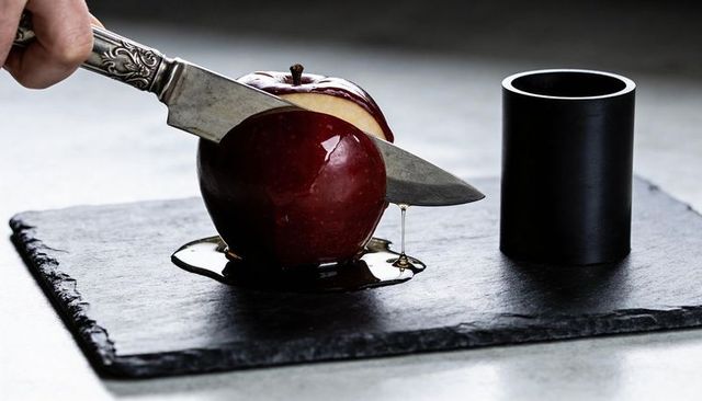 Slicing glossy red apple with ornate knife, honey dripping onto black slate platter close-up
