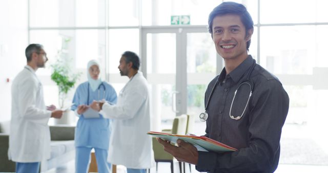 A smiling doctor holding medical records in a modern hospital environment. In the background, other healthcare professionals, including those from diverse ethnic backgrounds, are discussing medical information. Use for medical, healthcare, or hospital promotional material, representing teamwork in healthcare settings or illustrating a modern and diverse medical staff.