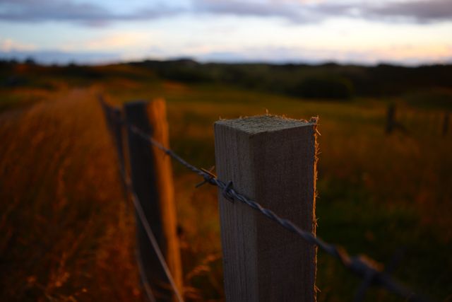 Barred fence illuminated by warm sunlight of dusk in countryside landscape. Ideal for conveying peaceful, rural emotions or backgrounds. Use in nature and outdoor scenes, background images, countryside tourism promotions, or for illustrating tranquility.