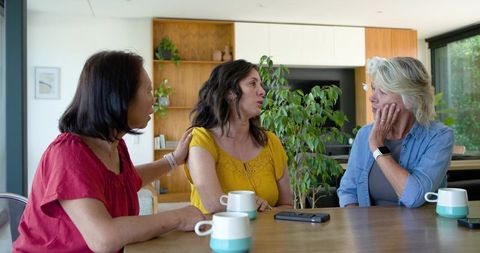 Multigenerational women sharing supportive conversation around kitchen table with mugs
