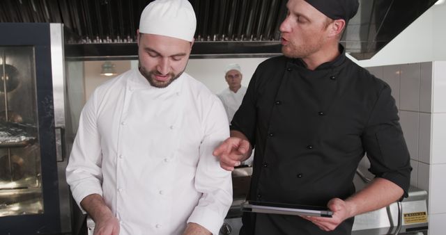 Chefs collaborating in a commercial kitchen, dressed in professional attire. One chef provides instructions while holding a tablet, the other chef listens attentively while cutting ingredients. Use for content related to culinary arts, professional cooking, teamwork in the kitchen, food preparation, and restaurant management.