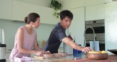 Couple preparing healthy breakfast at modern kitchen island, reaching for fruit bowl