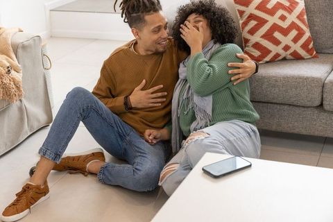 African american couple laughing and relaxing on living room floor with smartphone