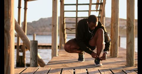african american man in sportswear tying his shoelace on boardwalk by ocean while listening to music through earphones. sunlight and ocean in background create motivational and energetic atmosphere. great for promoting healthy lifestyles, fitness programs, and sports gear.