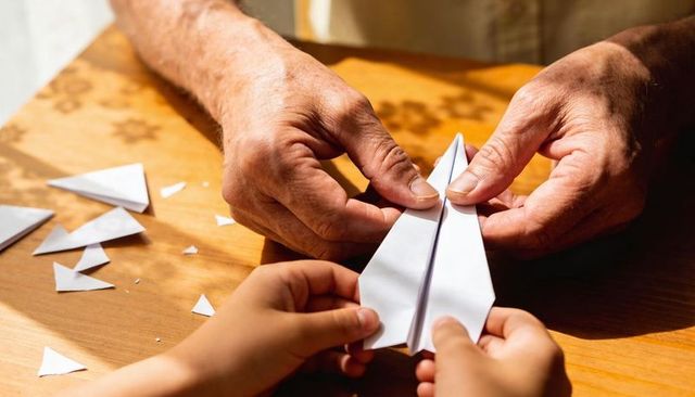Intergenerational guidance folding paper plane on sunlit wooden table with paper scraps