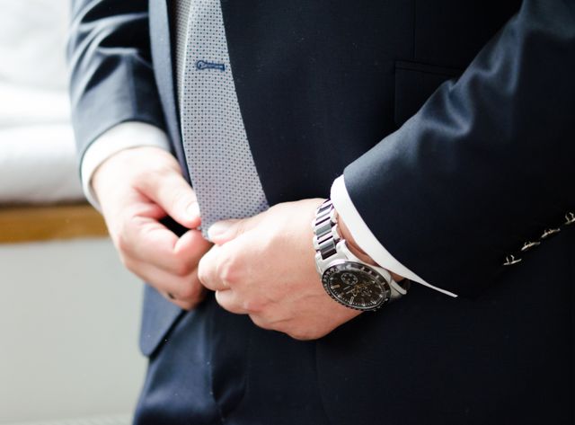 This image is perfect for websites, blogs, and advertisements focusing on business, professionalism, and corporate attire. It can be used to illustrate topics related to business etiquette, fashion, time management, or executive lifestyle. The emphasis on the hands adjusts a formal tie and suit, complemented by an elegant wristwatch, which conveys attention to detail and sophistication.