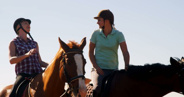 Two middle-aged Caucasian women enjoy a conversation while horseback riding under a clear blue sky, with copy space. Their casual attire and helmets suggest they are engaging in a leisurely equestrian activity.