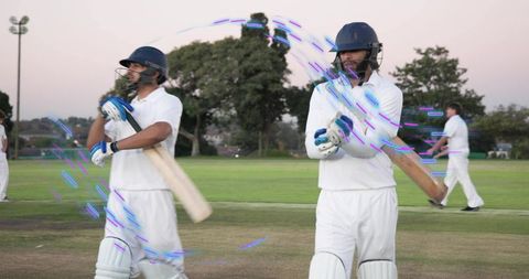 Two batsmen walking off pitch at twilight wearing white cricket gear on suburban ground