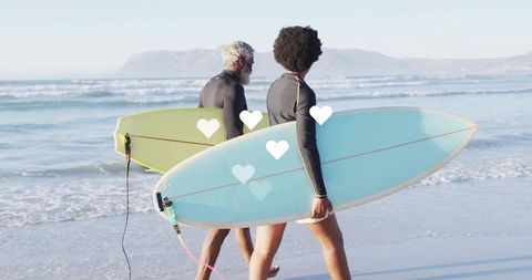 Two surfers walking along sunny shoreline carrying colorful surfboards in wetsuits