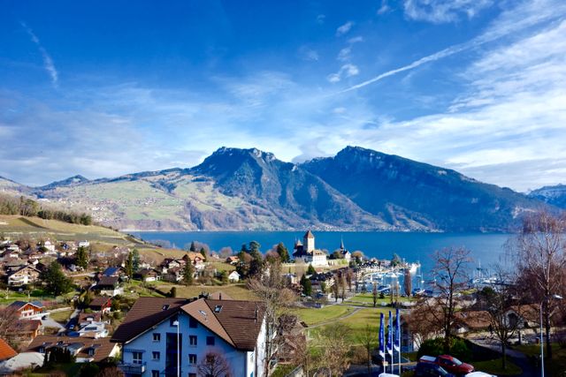 Photo captures an idyllic Swiss village situated by a calm lake, framed by majestic mountains. Houses and buildings spread across the landscape, creating a picturesque view under clear blue skies. Ideal for travel brochures, tourism promotions, scenery appreciation, desktop backgrounds, or nature and vacation-related content.