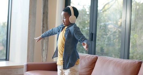 Joyful african american boy dancing on sofa wearing headphones in bright living room