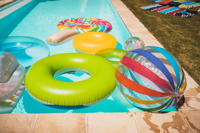 Colorful inflatable toys, including rings and a beach ball, floating in a garden pool on a sunny day. Ideal for use in advertisements for summer activities, pool parties, outdoor leisure products, and vacation promotions. Perfect for illustrating fun and relaxation during warm weather.