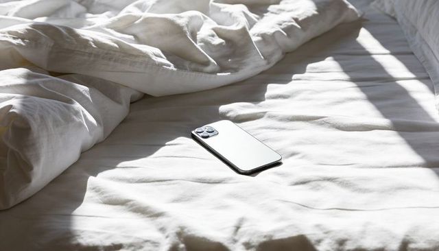 Modern smartphone resting on unmade white bed with morning sunlight and shadow bands