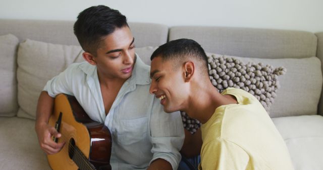 Young men relaxing on sofa, one playing guitar while the other is listening and smiling. Ideal for use in lifestyle blogs, friendship and relationship segments, music-related content, casual home settings, and social media campaigns focusing on young adult life and hobbies.