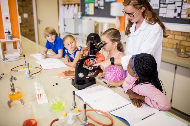 Teacher demonstrating microscope to a group of curious students in a science classroom. Children are gathered around the teacher, showing interest in the experiment. Various lab equipment and colorful liquids are visible on the table. Ideal for educational content, science promotion, and classroom activities.