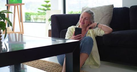 Mature woman relaxing on floor by sofa checking smartphone casual living room lifestyle