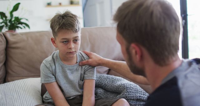 Father gently comforts and supports his sad son who is sitting on a couch. The interaction highlights themes of empathy, care, and parenting in a home environment. This can be used in articles or content related to mental health, emotional support, family dynamics, and parental involvement.