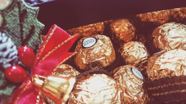 Up-close image showing several Ferrero Rocher chocolates wrapped in golden foil, surrounded by festive holiday decor featuring a Christmas tree, ribbon, and bell. Perfect for holiday-themed marketing, advertisements, greeting cards, or social media campaigns celebrating the festive season and gift-giving. Ideal to evoke feelings of luxury and indulgence.