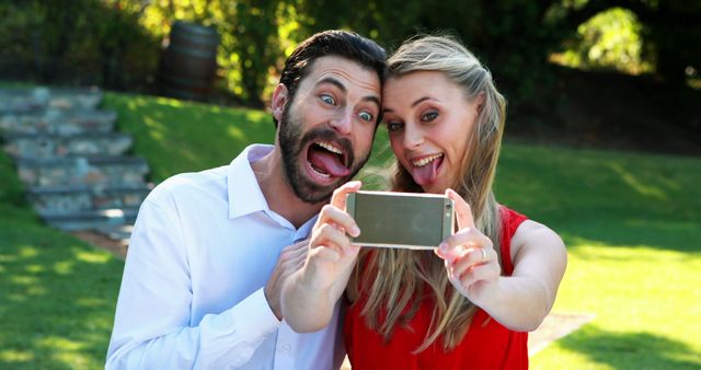 Couple enjoying a bright sunny day while taking a playful selfie in a green park. Ideal for themes of togetherness, fun, outdoor activities, social media, and carefree moments. Suitable for websites, social media posts, and blogs promoting relationships, leisure, and technology use.