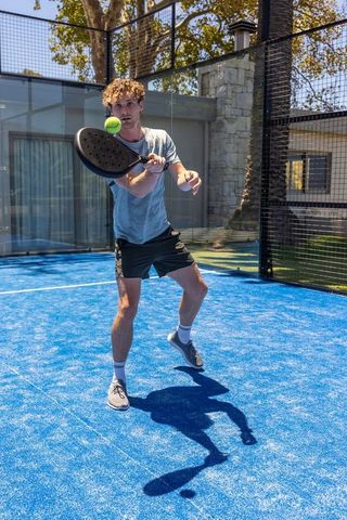 Young man playing padel on bright blue court hitting forehand with black paddle and ball