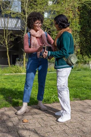 Female friends exchanging tablet on college campus walkway wearing scarves and backpacks