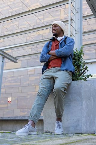Young african american man relaxing on urban planter wearing headphones, beanie and backpack