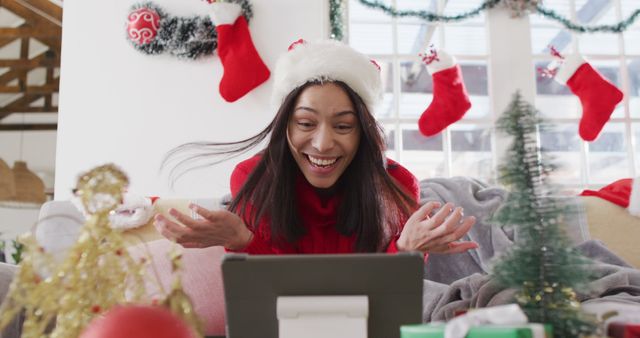 Cheerful woman wearing a Santa hat video calling during Christmas celebration, surrounded by festive decorations. Ideal for holiday greeting cards, Christmas advertisements, virtual communication, and social media campaigns emphasizing holiday cheer and long-distance connections.