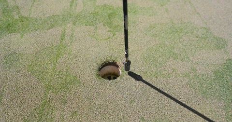 Flagstick casting diagonal shadow over golf hole on topdressed putting green closeup