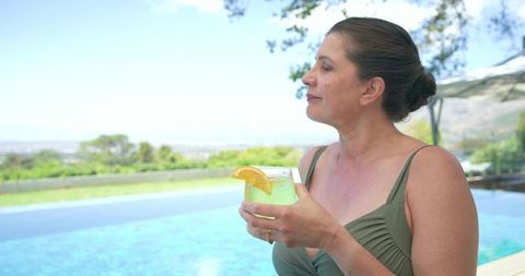 Mature woman relaxing poolside sipping citrus cocktail with scenic garden and ocean view
